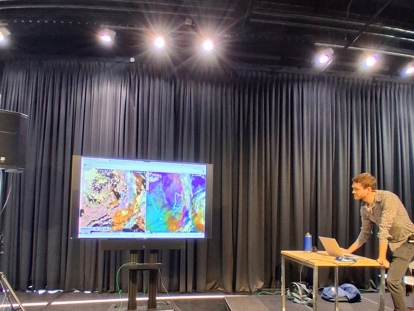 A speaker at a laptop on a stage beside a screen showing weather maps
